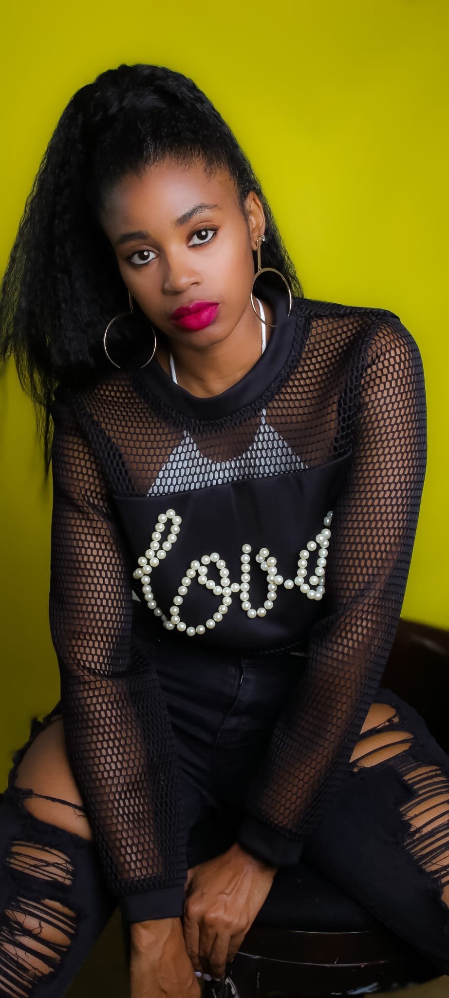 Lovett Love seated in a black mesh top and ripped jeans against a chartreuse wall
